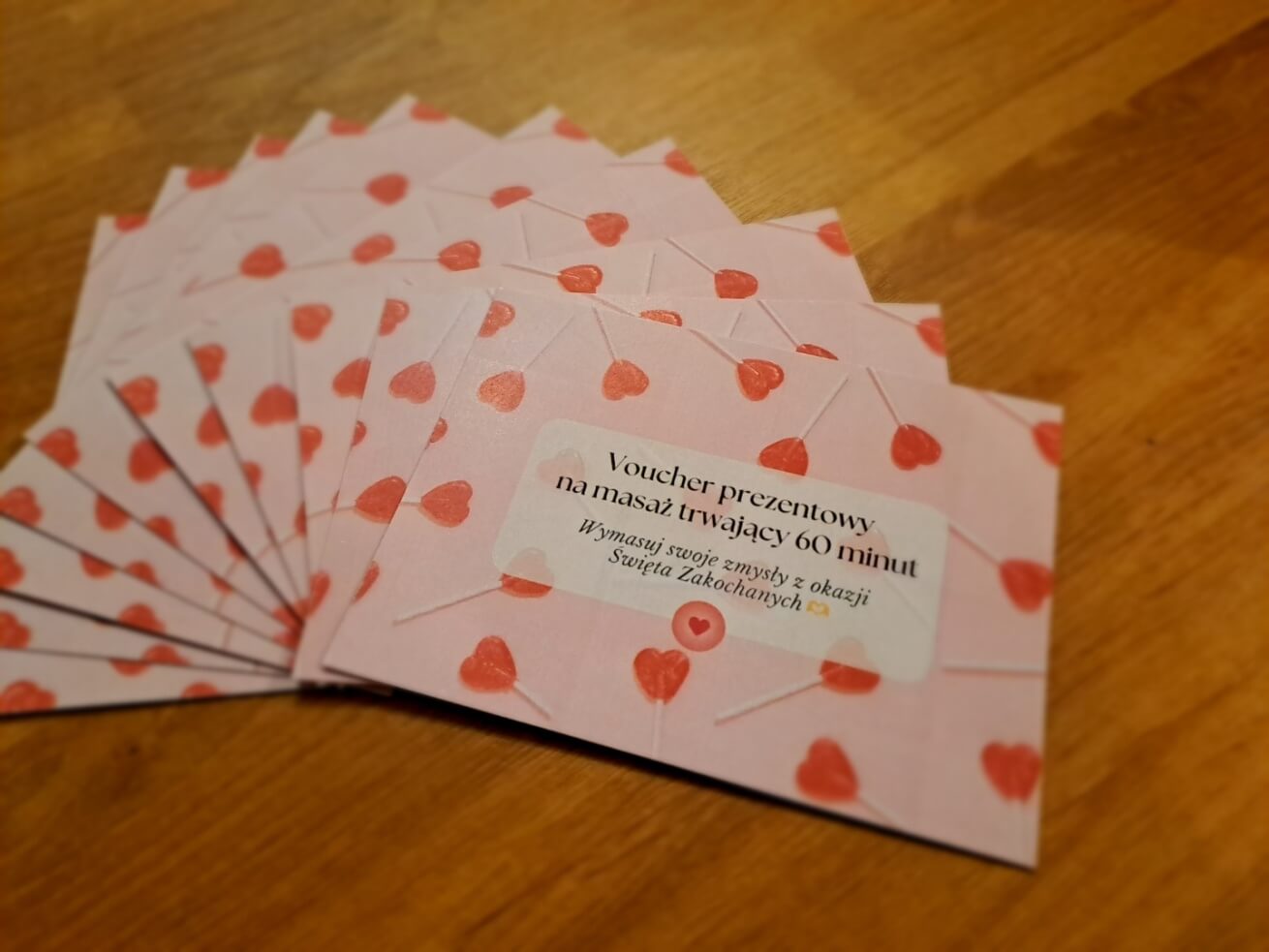 Wachlarz różowych walentynkowych voucherów prezentowych na masaż, ozdobionych czerwonymi lizakami w kształcie serc. Na pierwszym planie voucher z napisem 'Voucher prezentowy na masaż trwający 60 minut' i podtekstem 'Wymasuj swoje zmysły z okazji Święta Zakochanych'.